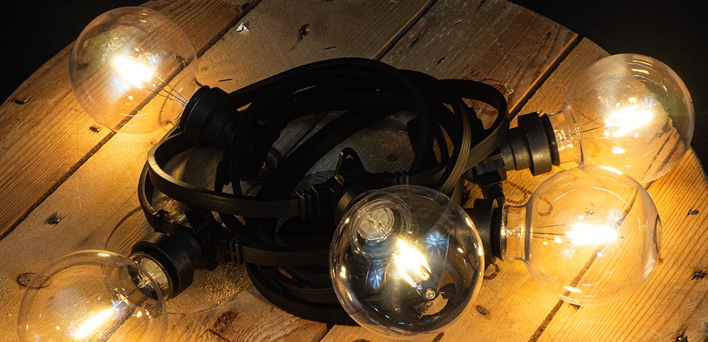 Dekoracyjna żarówka LED filament G95 1W z plastikowym kloszem, zawieszona w girlandzie zewnętrznej, emitująca ciepłe światło 2700K, nastrojowa iluminacja plenerowa o zmierzchu.