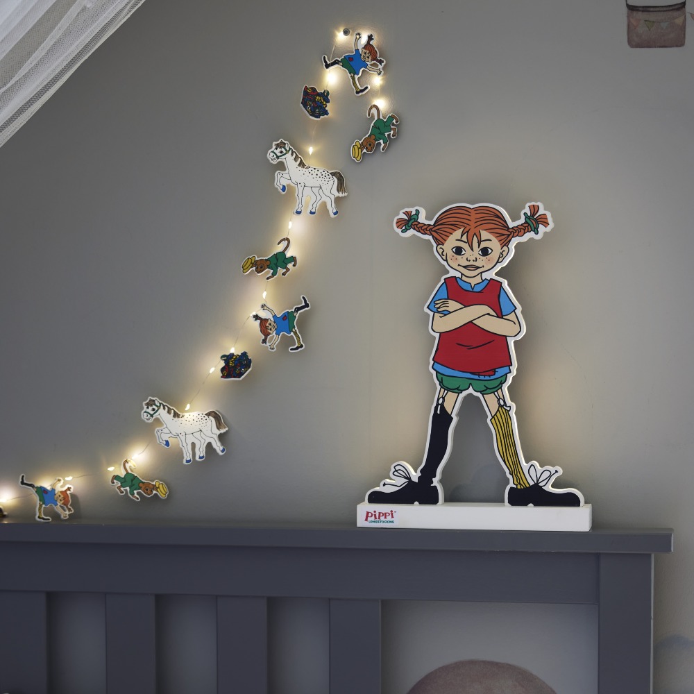 Podświetlana sylwetka Pippi Pończoszanki ustawiona na oparciu łóżka, w towarzystwie dekoracyjnej girlandy świetlnej, tworzy przytulny i bajkowy klimat w pokoju dziecka.