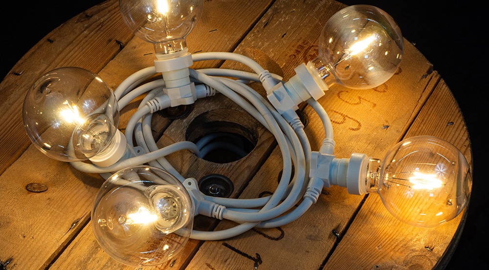 Dekoracyjna żarówka LED filament G95 1W z plastikowym kloszem, zawieszona w girlandzie zewnętrznej, emitująca ciepłe światło 2700K, nastrojowa iluminacja plenerowa o zmierzchu.