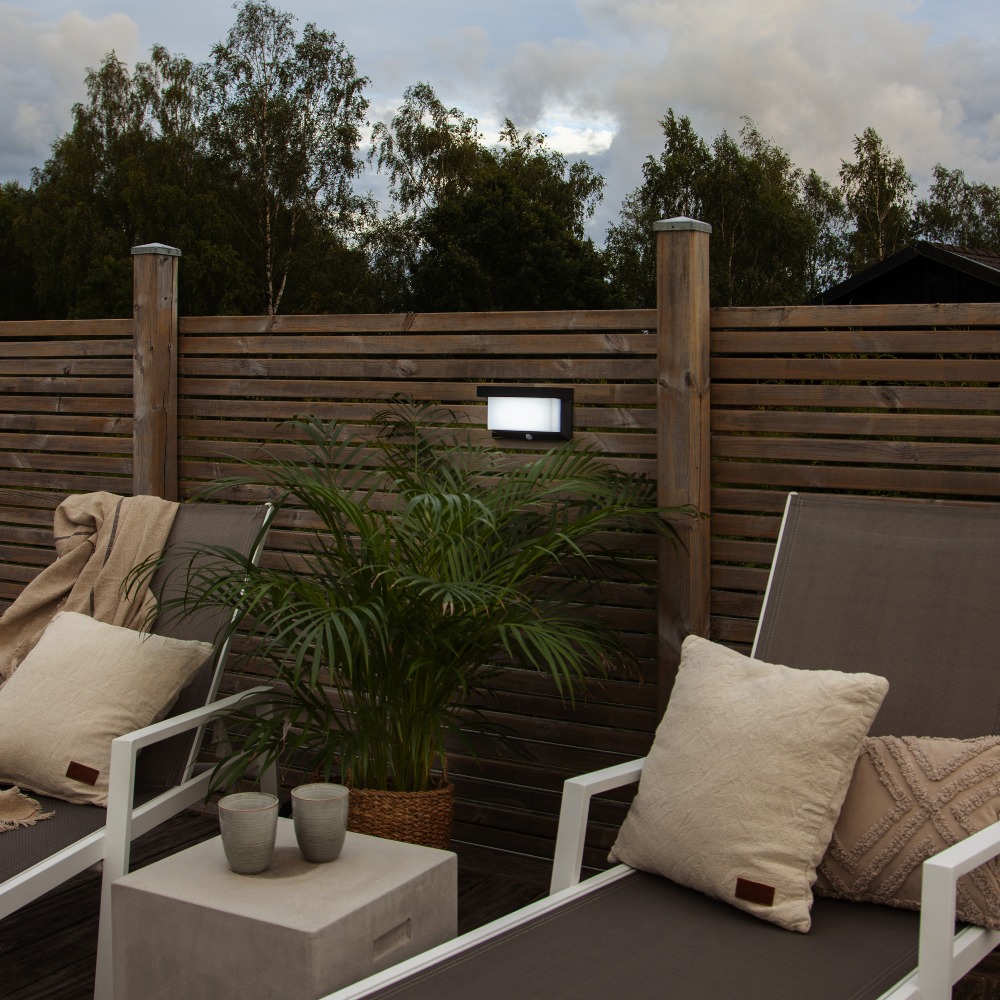 Kinkiet solarny zamontowany na ogrodzeniu tarasu, emituje białe światło i tworzy przytulną atmosferę w strefie wypoczynkowej z roślinami i meblami ogrodowymi.