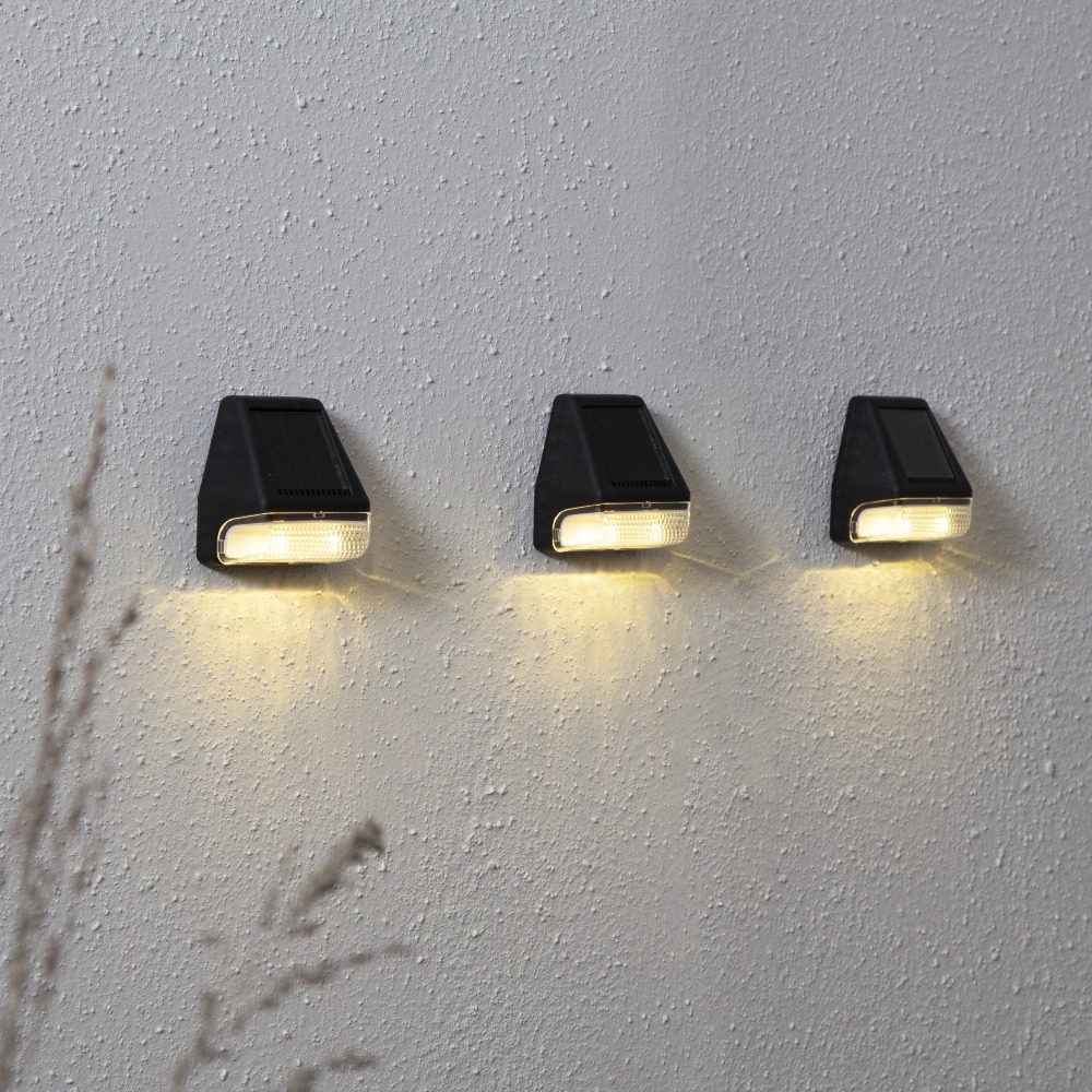 Trzy lampy solarne LED zamontowane na ścianie, emitujące ciepłe światło skierowane w dół, idealne do oświetlenia elewacji lub ogrodu.