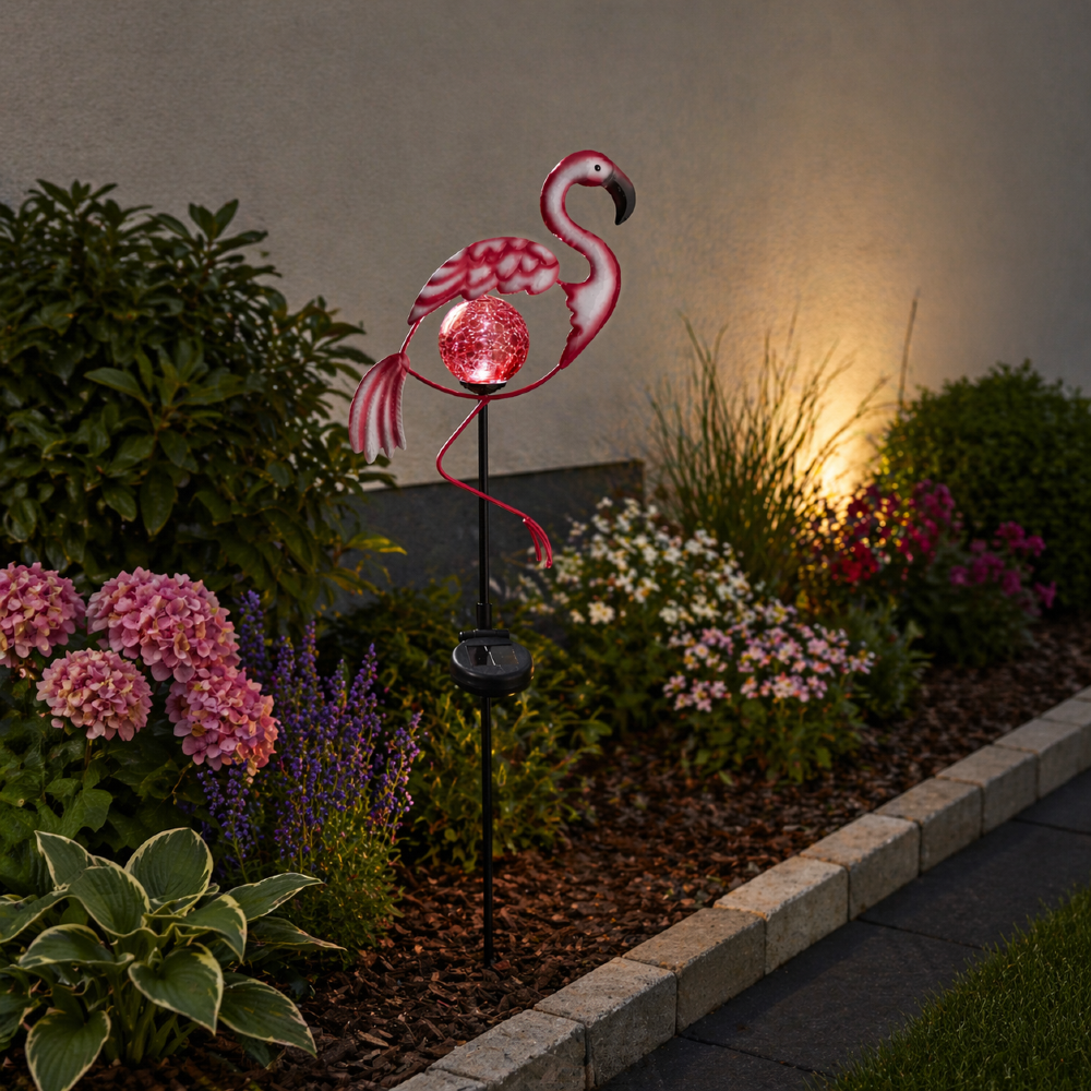 Solarna dekoracja ogrodowa flaming z podświetlaną kulą, ustawiona wśród kwiatów przy rabacie, subtelnie rozświetla przestrzeń i dodaje jej dekoracyjnego charakteru po zmroku.