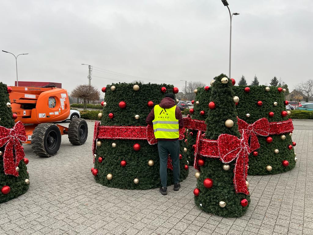 Montaż dekoracyjnej choinki modułowej z czerwonymi kokardami i bombkami. Na placu leżą sekcje choinki, obok pracownik w kamizelce z logo AMLED i podnośnik koszowy. Tło stanowi parking z lampami i pochmurne niebo.