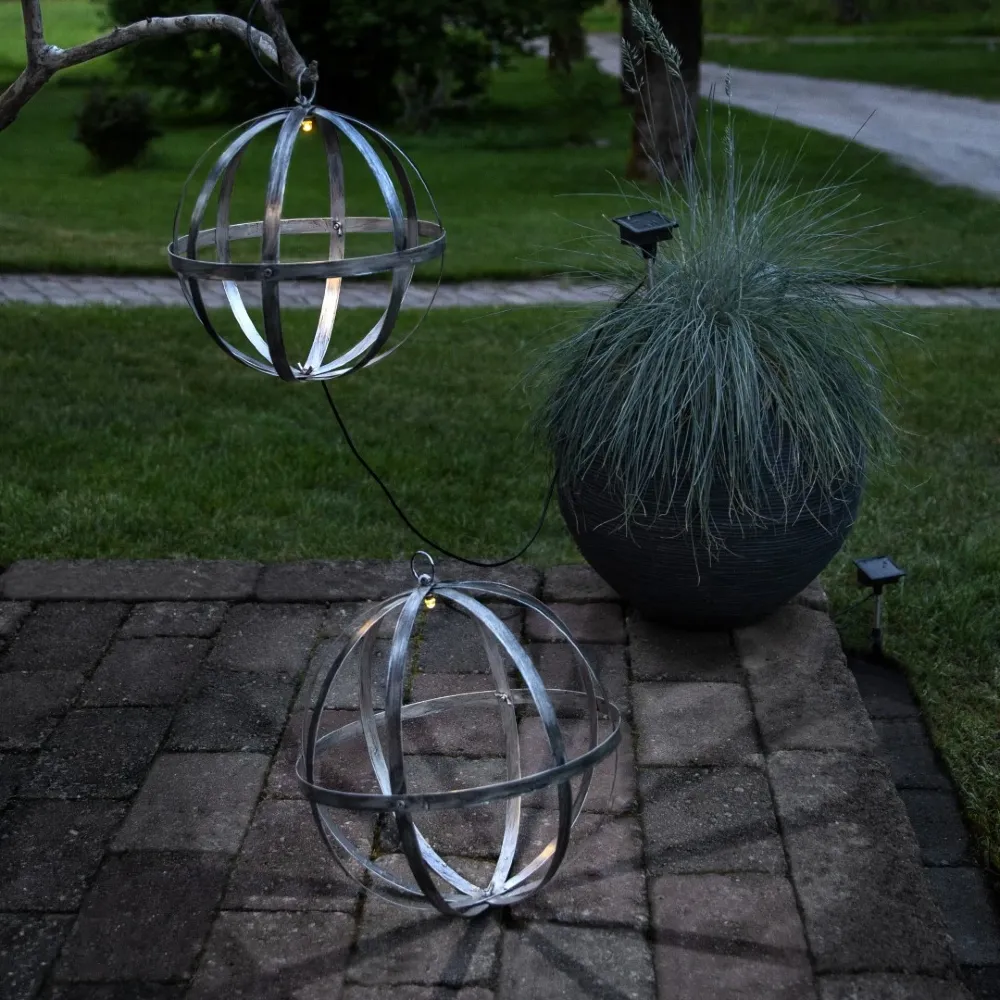 Dwie metalowe kule solarne LED wykorzystane jako dekoracja tarasu i ogrodu. Jedna kula ustawiona jest na kostce brukowej, druga zawieszona nad nią, a delikatne światło podkreśla geometryczny kształt obu ozdób.