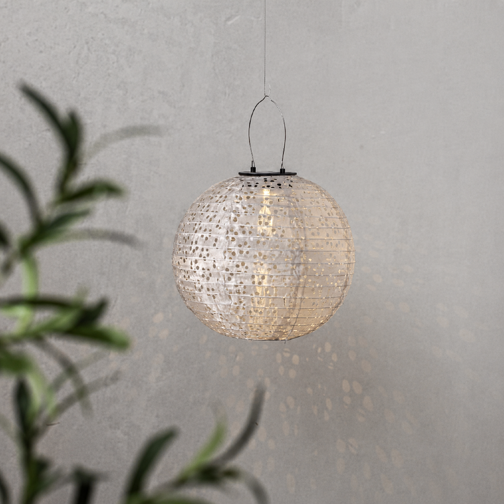 Wisząca lampionowa kula solarna z ażurowym wzorem emituje ciepłe światło, tworząc dekoracyjne refleksy na jasnej ścianie w tle.