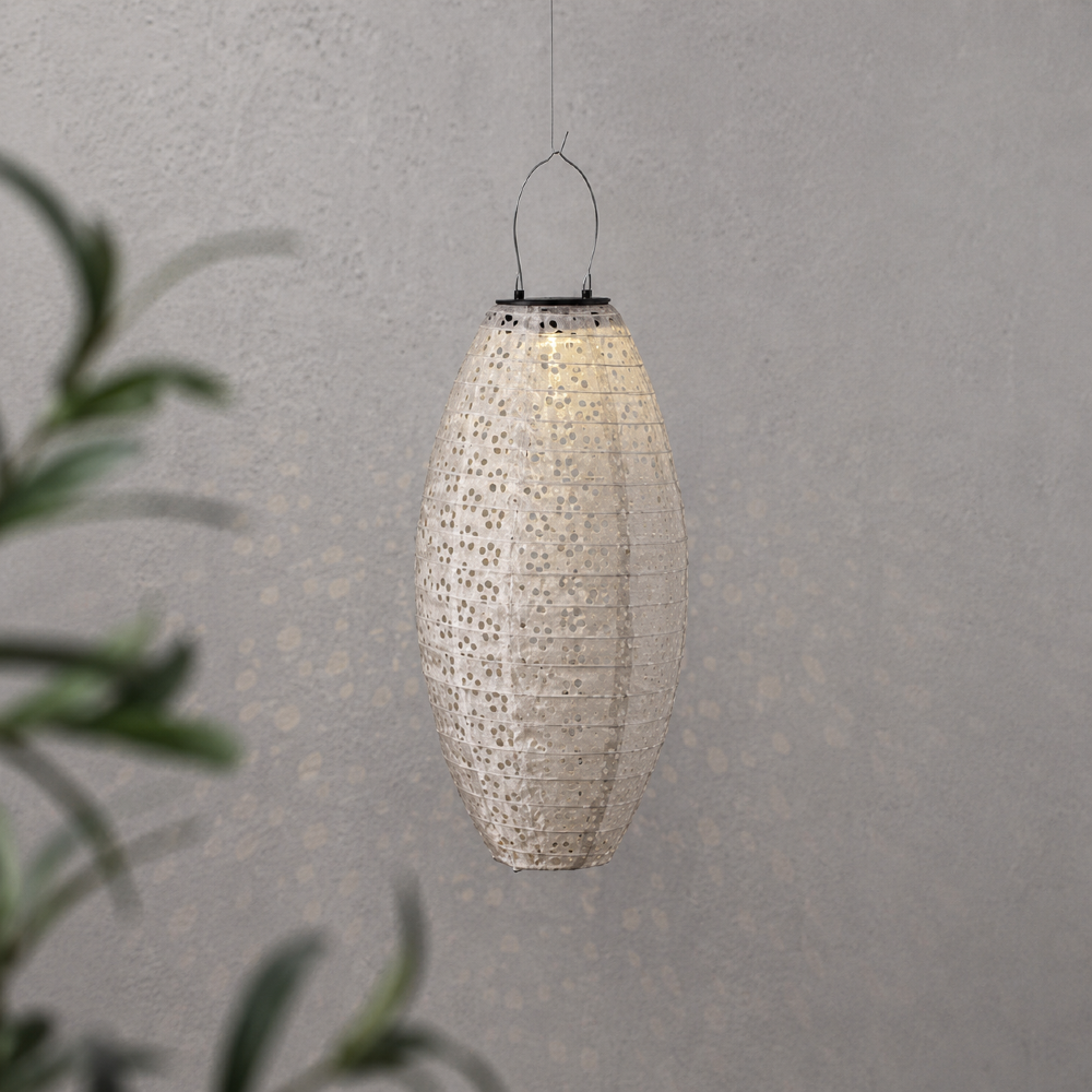 Ażurowy lampion solarny LED o podłużnym kształcie zawieszony na cienkiej lince. Delikatnie rozprasza światło, tworząc subtelne refleksy na jasnej ścianie.
