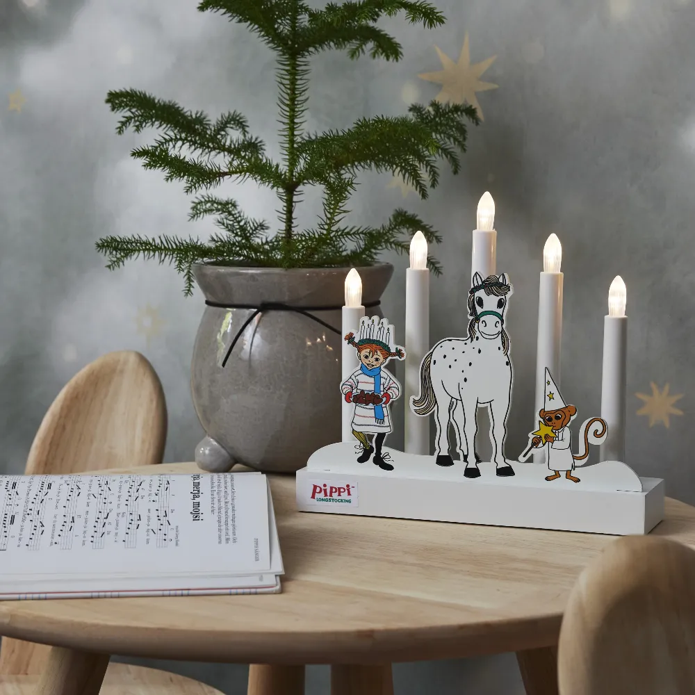 Świecznik LED z bohaterami Pippi Pończoszanki ustawiony na stole obok otwartej książki, doniczek z roślinami i zapalonych świec, tworząc przytulną świąteczną aranżację.