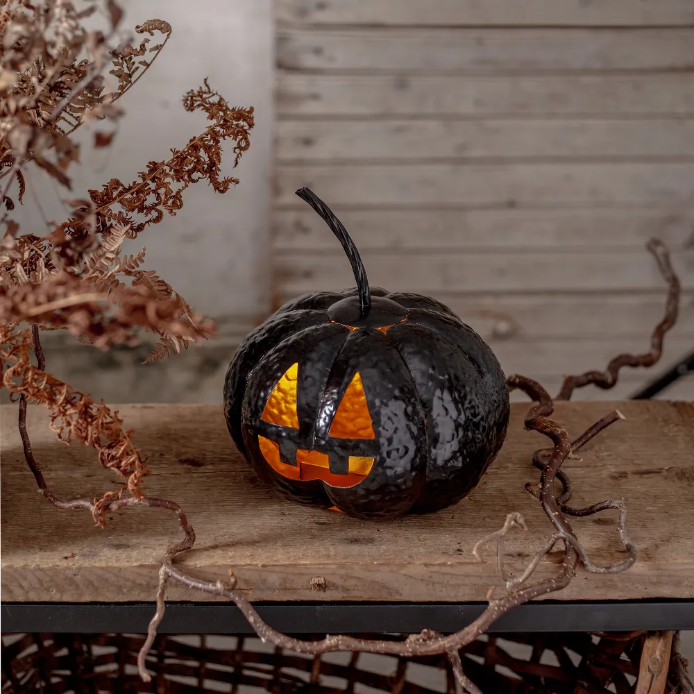 Świecąca czarna dynia Halloween ustawiona na drewnianej półce w aranżacji z suchymi liśćmi, gałązkami i jasnym tłem.