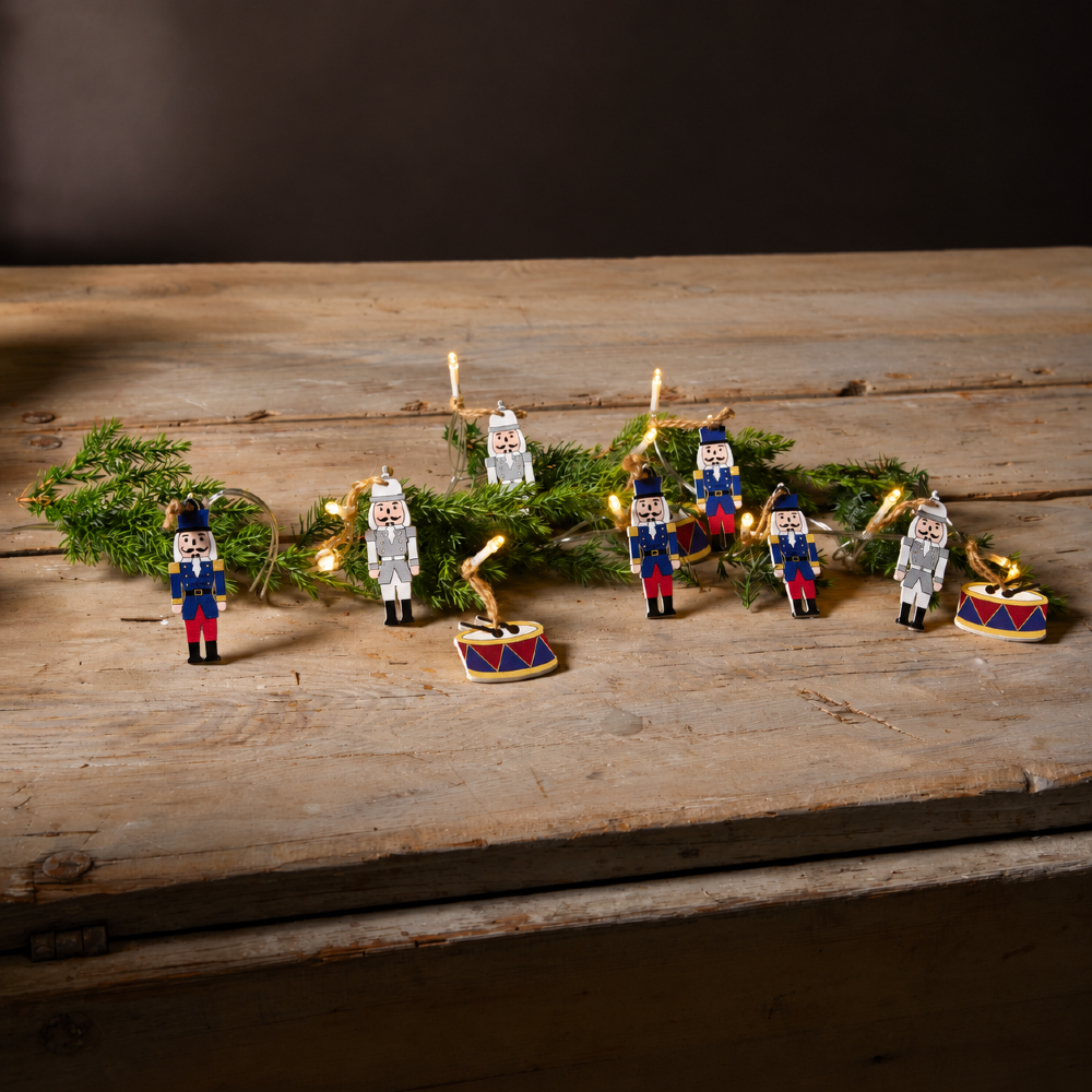Łańcuch świąteczny LED z drewnianymi figurkami dziadków do orzechów w granatowych i szarych mundurach oraz kolorowymi bębenkami, ułożony wśród zielonych gałązek na rustykalnej drewnianej skrzyni.