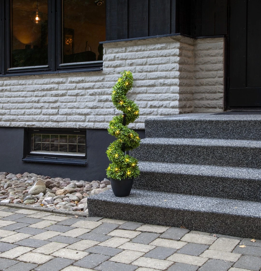 Spiralna dekoracja bukszpanowa z oświetleniem LED ustawiona przy wejściu do domu, na tle schodów i elewacji.