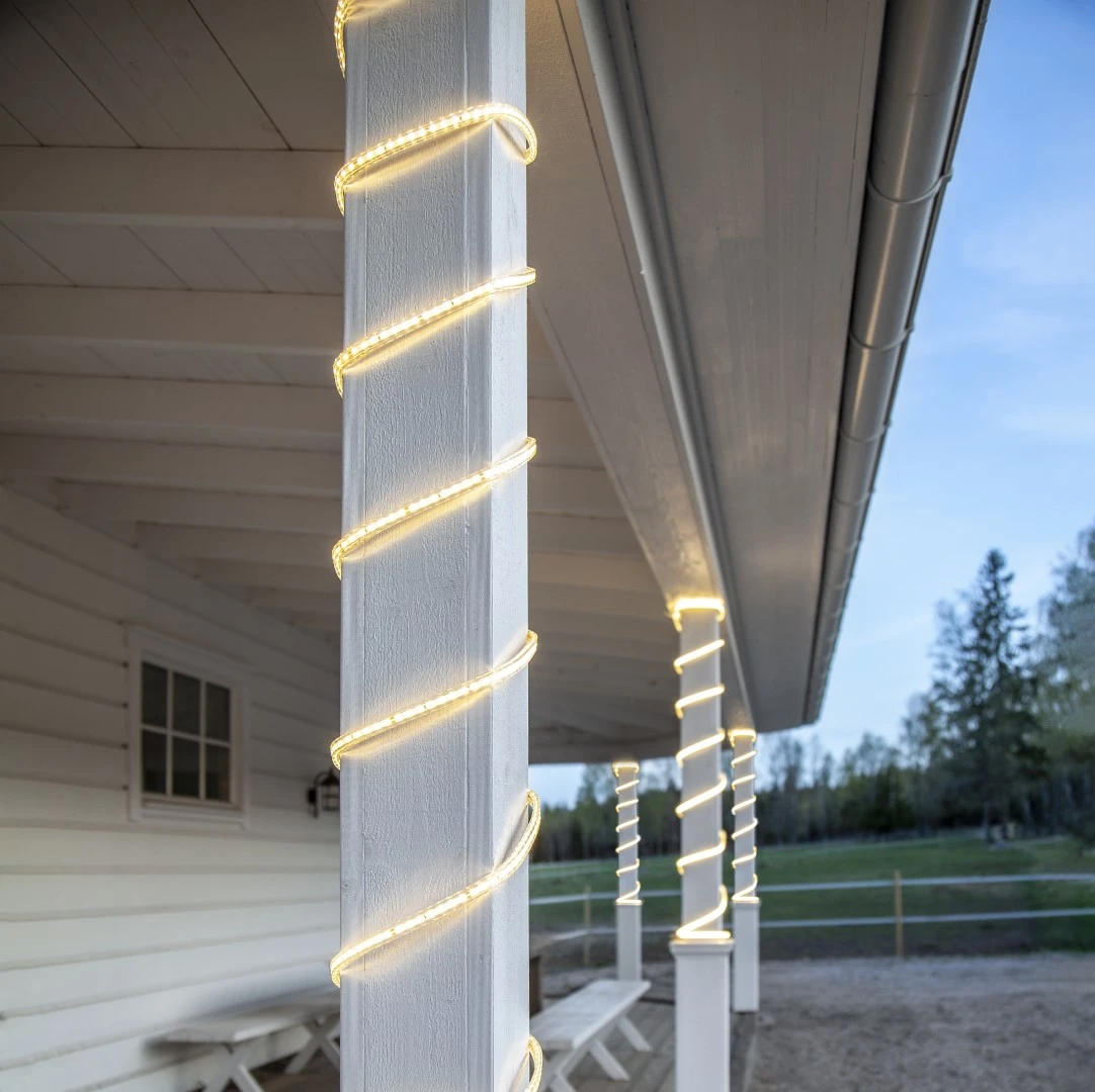 Wąż LED owinięty spiralnie wokół białych drewnianych kolumn na ganku domu, tworzący dekoracyjne oświetlenie.